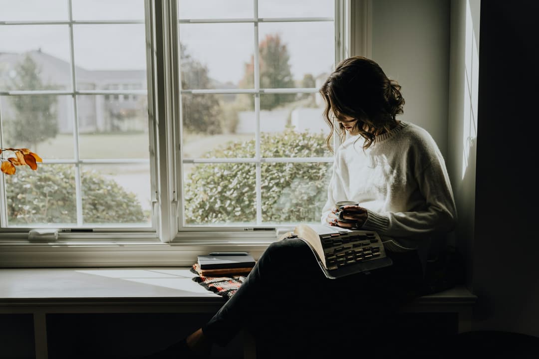 Vrouw leest bij het raam, omringd door boeken
